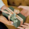 Two hands exchanging an elegantly wrapped green gift box with ribbons and foliage., tags: bleibende eindrücke - pexel