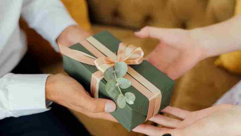 Two hands exchanging an elegantly wrapped green gift box with ribbons and foliage., tags: bleibende eindrücke - pexel